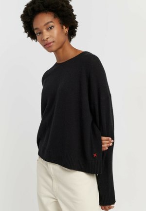 Zwarte gebreide trui met ronde hals, doorgezakte schouders en geribbelde zoom. Heeft een klein rood X-accent aan de linkerkant.