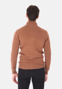 Maglione marrone con colletto alto e orlo a coste, realizzato in un tessuto morbido. Il modello indossa pantaloni neri; ripreso da dietro su uno sfondo bianco.