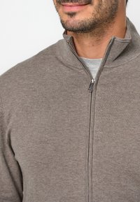 Maglione marrone con zip, colletto alto, tessuto lavorato e dettaglio della zip in argento, indossato sopra una maglietta grigia.