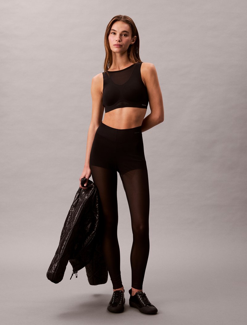 Soutien desportivo preto com detalhes em rede, leggings pretas de cintura alta e uma jaqueta preta acolchoada segurada numa mão. Ténis pretos completam o visual.