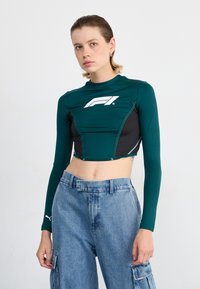 Groene en zwarte crop top van stof met lange mouwen; heeft een wit logo op de borst en contrasterende stiksels. Gepaard met losse denim cargobroek.