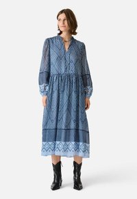 Sininen kuviollinen mekko, jossa on puoliksi läpinäkyvä kangas, kaulus, pitkät hihat ja kerätty vyötärö. Yhdistetty mustiin nilkkasaappaisiin.