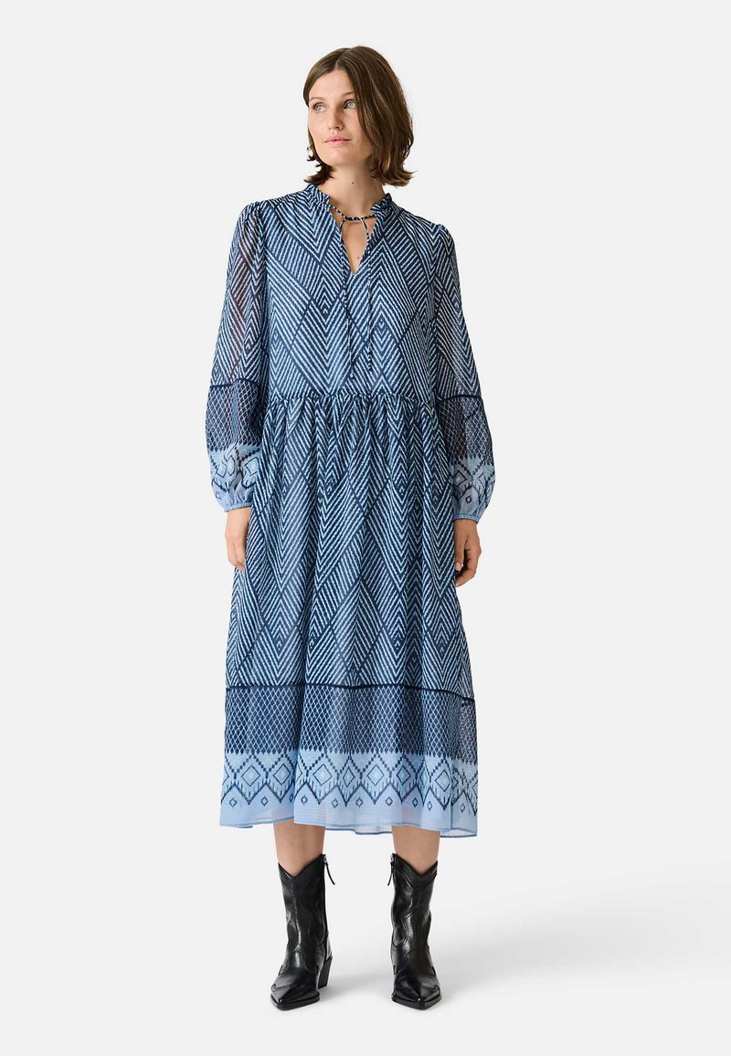Sininen kuviollinen mekko, jossa on puoliksi läpinäkyvä kangas, kaulus, pitkät hihat ja kerätty vyötärö. Yhdistetty mustiin nilkkasaappaisiin.