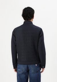 Giacca marina con retro trapuntato e maniche lisce, dotata di colletto alto e polsini con bottoni. Indossata sopra jeans in denim blu chiaro.