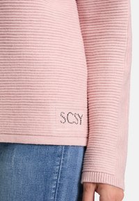 Jasnoróżowy sweter prążkowany z długimi rękawami, z napisem "SCSY" wysadzanym cyrkoniami na dolnym obszyciu. Miękka tekstura, casualowy design.