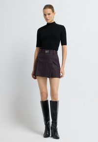 Kvinde iført en sort kortærmet turtleneck, mørk plisseret miniskørt med bælte og knæhøje sorte støvler foran en ensfarvet baggrund.