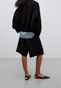 Svart oversized tröja, ljusblå skjorta under, svarta shorts och svart spetsiga flats. Matta ytor med minimal detaljering.