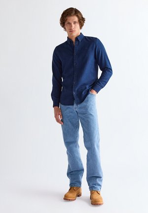 Jeune homme debout, portant une chemise bleu foncé boutonnée, un jean bleu clair et des bottes de travail beige, sur un fond blanc uni.