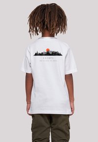 Child with brown dreadlocks wearing white T-shirt with black and red mountain and building graphic and the words "ESCAPE AND DISCOVER THE WORLD" on back.