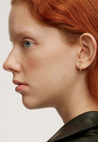 Pendiente de aro dorado con diseño de tipo rama, colocado contra una oreja. Superficie lisa, pequeñas cuentas acentúan las ramas. Textura natural de la piel visible.