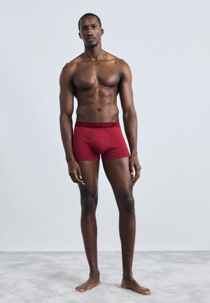 Röda kalsonger i boxermodell med logoband, figursydd design, tillverkade av ett glatt material, visade på en muskulös manlig modell mot en neutral bakgrund.