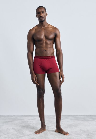 Boxeri roșii cu talie logo, design ajustat, fabricați dintr-un material neted, expuși pe un model masculin muscular pe un fundal simplu.