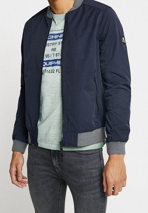 Mand iført en navy bomberjakke med grå manchetter over en lysegrøn T-shirt med grafik og grå jeans.