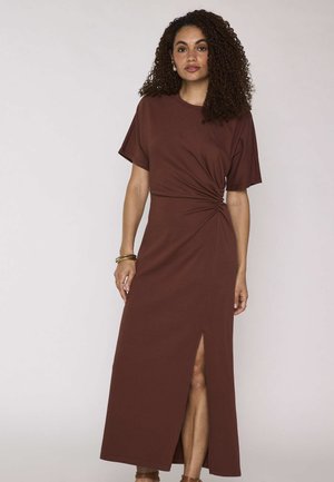 Vrouw met krullend haar die een bruine maxi-jurk met korte mouwen, zijsplooien en een voorsplit draagt, geaccentueerd met armbanden en oorbellen.