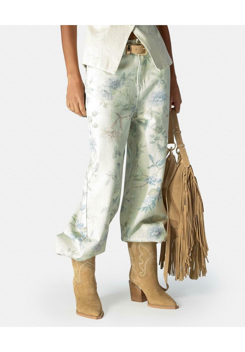 Personne portant un pantalon léger à fleurs et des bottes de cowboy en daim marron clair, tenant un sac à main en daim marron avec des franges et des sangles à boucle.