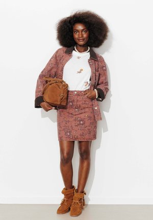 Femme avec un grand afro portant un ensemble veste et jupe à fleurs roses, un t-shirt blanc à fleurs, des bottes en daim marron, et tenant un sac en daim marron.