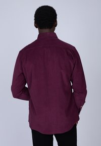 Chemise en velours côtelé bordeaux à manches longues, avec un col et un ourlet arrondi. Texture visible et détail de boutons aux poignets.