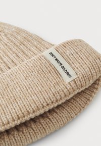 Beige kootud beanie ribakujulise tekstuuriga, millel on kangasilt, kus on kirjas "ÄRA RAASTA KULTUURI." Minimaalne ja soe disain.