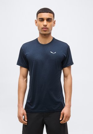 Νέος άνδρας που στέκεται προσανατολισμένος προς τα εμπρός, φορώντας απλό μπλε σκούρο κοντομάνικο πουκάμισο με μικρό λευκό λογότυπο στο στήθος.