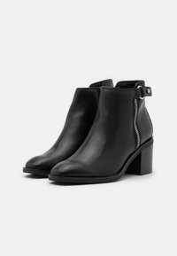 Botas de cuero negro hasta el tobillo con punta redondeada, cierre lateral con cremallera y un tacón cuadrado robusto. Presentan una correa decorativa ajustable.