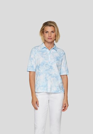 Helles blaues Poloshirt mit floralem Muster, kurzen Ärmeln, Knopfleiste und einem Logodetail am Saum. Kombiniert mit weißen Hosen.