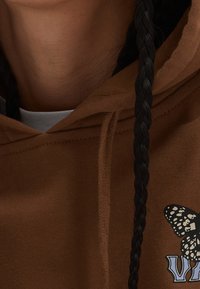 Brown hoodie with a drawstring, featuring a butterfly graphic and the word "VAN" in blue. The fabric appears soft with a textured finish.