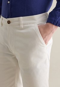 Weiße Baumwollhosen mit schlankem Schnitt, ausgestattet mit Seitentaschen und einem einzelnen braunen Knopf an der Taille, kombiniert mit einem blauen Hemd mit Knöpfen.