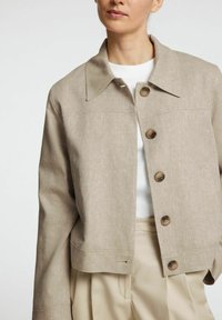 Chaqueta de lino beige claro con un collar en punta, botones en el frente y dos bolsillos en el pecho, combinada con una camisa blanca y pantalones beige.