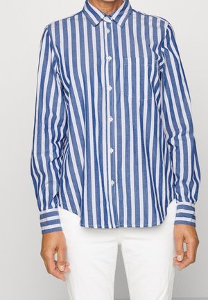 Person wearing a blue and white vertically striped button-up shirt with a chest pocket and white pants, standing with arms relaxed.