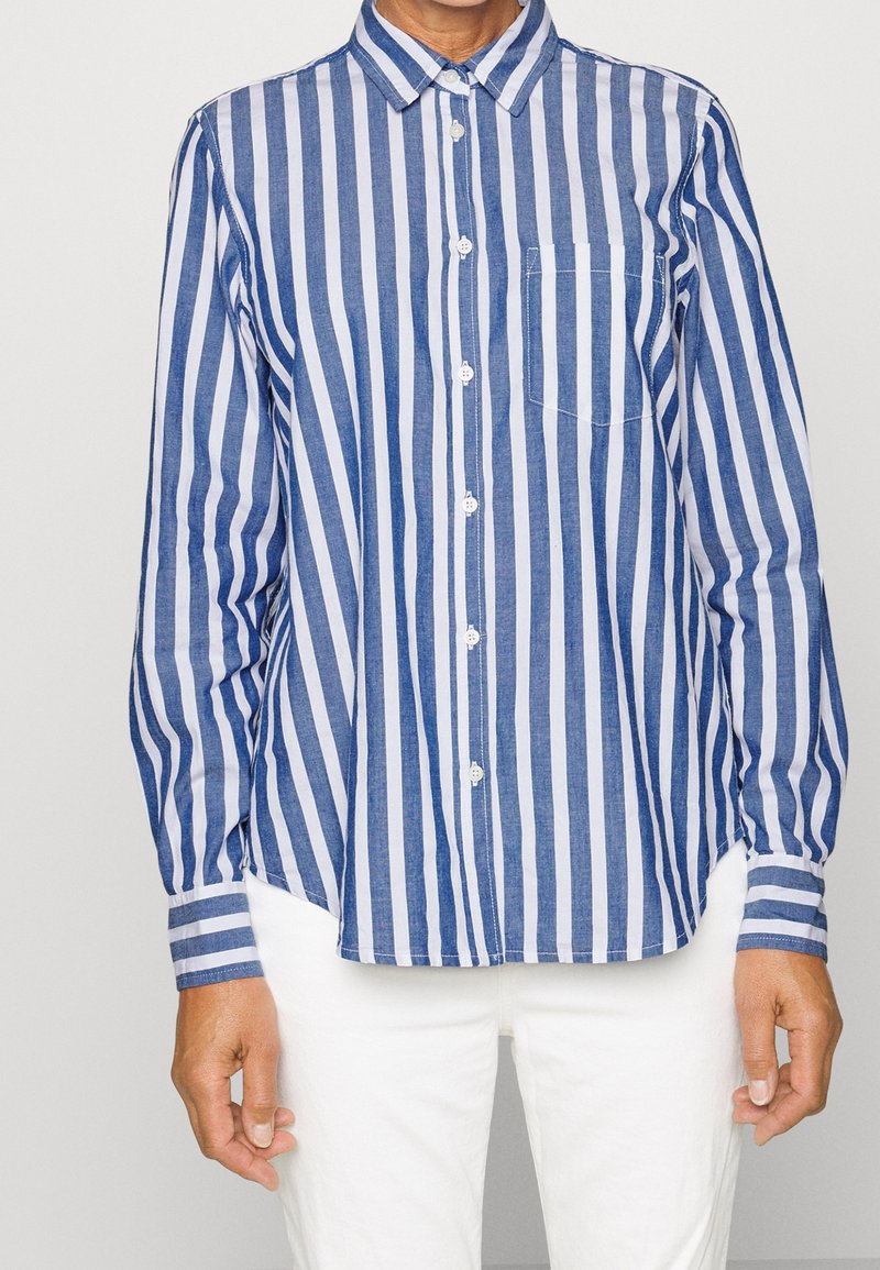 Personne portant une chemise à rayures verticales bleues et blanches avec une poche poitrine et un pantalon blanc, debout, les bras détendus.