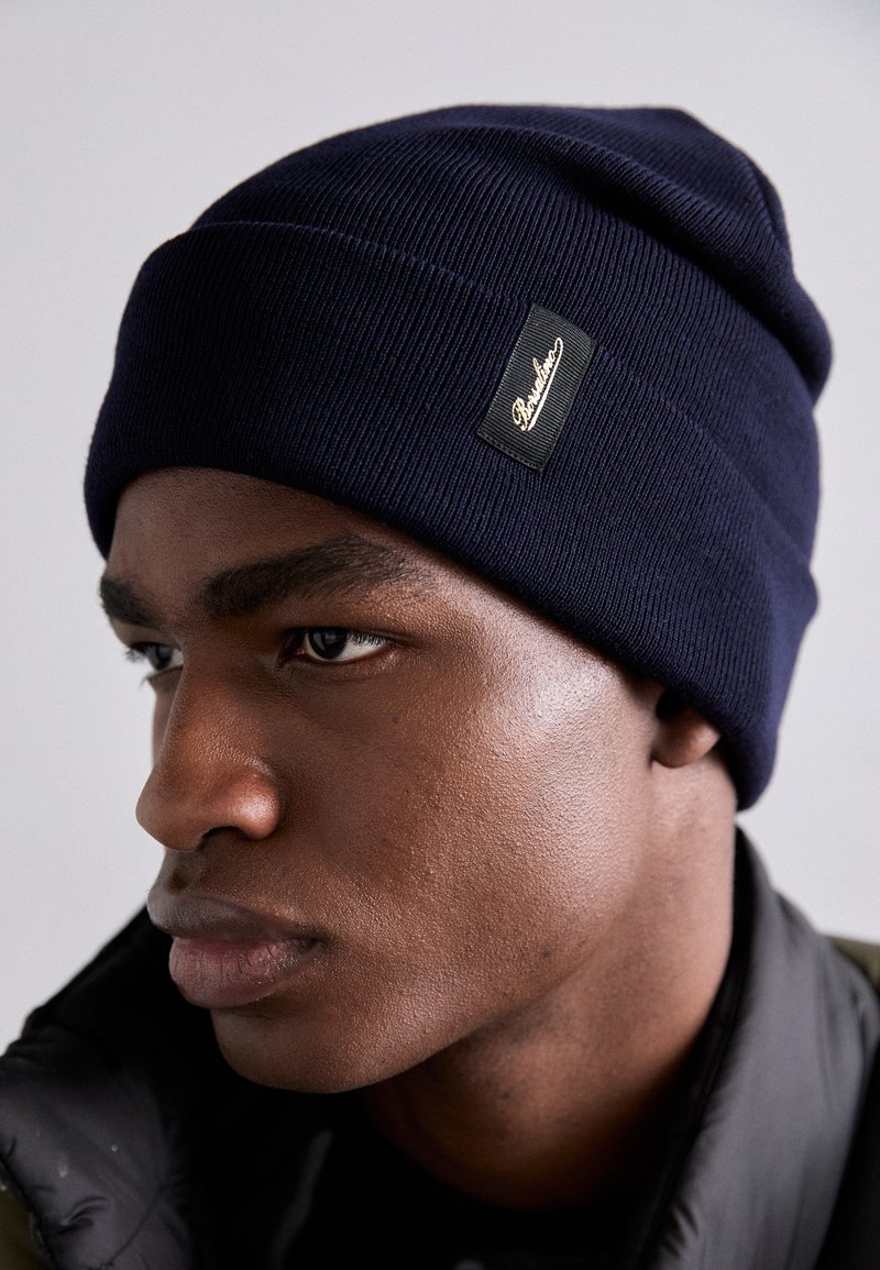 Tamsiai mėlynas įsukamas megztas skrybėlis, kuriame yra juodas etiketė. Dizainas yra prigludęs, su aptempta viršutine dalimi ir sukta kraštu šilumai.