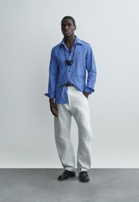 Homme debout, une main dans la poche, portant une chemise à rayures bleues, un pantalon blanc à jambes larges, une ceinture noire, des chaussures noires et un masque autour du cou.