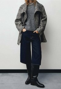Manteau en tweed gris sur un pull gris, associé à un jean large bleu foncé et des bottes noires montant jusqu'aux genoux. Accessoires minimalistes et arrière-plan neutre.