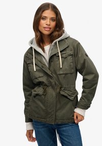 Chaqueta militar de color verde oliva con cierre de cremallera y botón, dos bolsillos en el pecho, cintura ajustable con cordón y capucha gris desmontable.