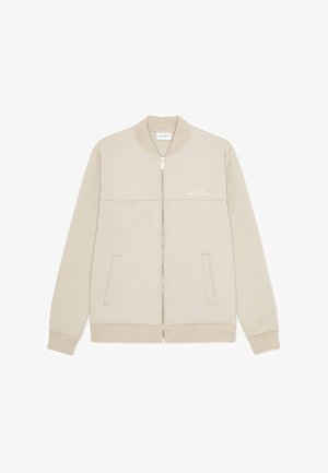 Beige bomberjakke med glidelås, med glatt tekstur, ribbestrikkede mansjetter, to frontlommer og en subtil logo på brystet.