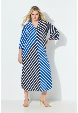 Kobieta ubrana w niebieską i czarną midi sukienkę w przekątne prążki z rękawami do łokcia i złote sandały na wysokim obcasie, stojąca przy białej ścianie.