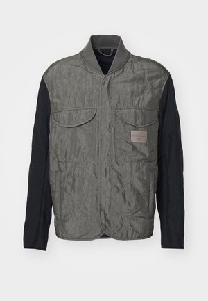 Gilet matelassé gris avec des manches noires, deux poches poitrine, col côtelé et une étiquette de logo sur le côté gauche. Tissu lisse et texturé.