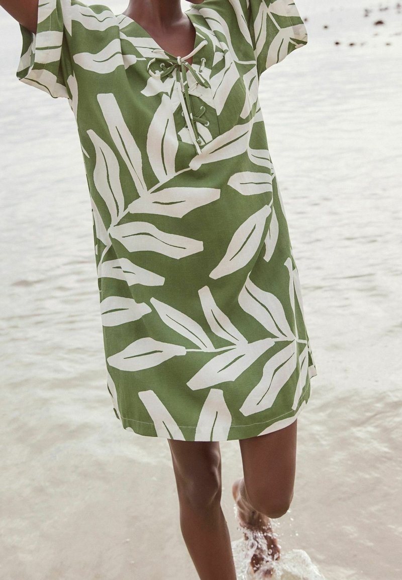 Personne portant une robe verte et blanche à motif de feuilles, debout avec un pied dans l'eau peu profonde à la plage.