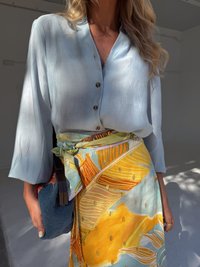 A woman wearing a light blue blouse and a colorful patterned skirt holds a stylish blue bag, standing in a well-lit indoor space.