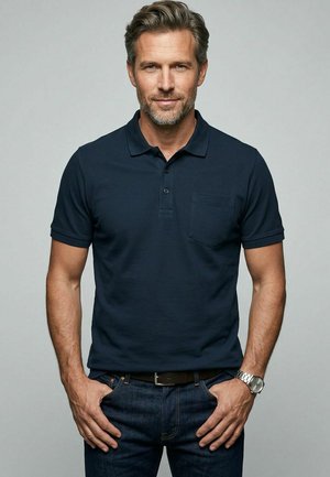 Mann mit hellbraunen Haaren und Bart trägt ein dunkelblaues Polohemd und Jeans, steht mit den Händen nahe den Taschen vor grauem Hintergrund.