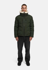 Žalia pūkinė striukė su dirbtinio kailio apykakle, turinti horizontalius siūlus, dvi užtrauktuku uždaromas kišenes ir priekinį užtrauktuką, derinama su juodomis velveto kelnėmis.