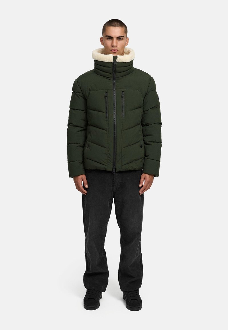 Žalia pūkinė striukė su dirbtinio kailio apykakle, turinti horizontalius siūlus, dvi užtrauktuku uždaromas kišenes ir priekinį užtrauktuką, derinama su juodomis velveto kelnėmis.