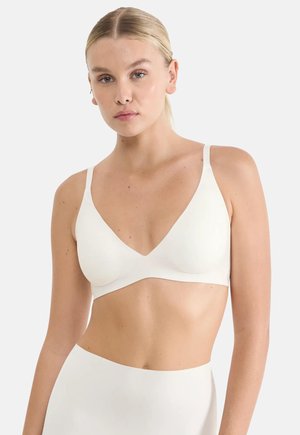 Žena s blond vlasy má na sobě bílou bezšvovou podprsenku bez kostic a odpovídající spodní díl s vysokým pasem na jednoduchém světlejším pozadí.