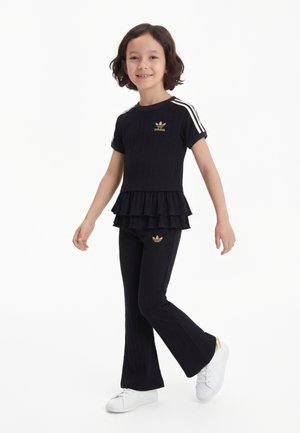 Młoda dziewczyna uśmiecha się, ubrana w czarny strój Adidas ze złotymi logo, białe paski na rękawach, czarny marszczony top, rozszerzane spodnie i białe trampki.