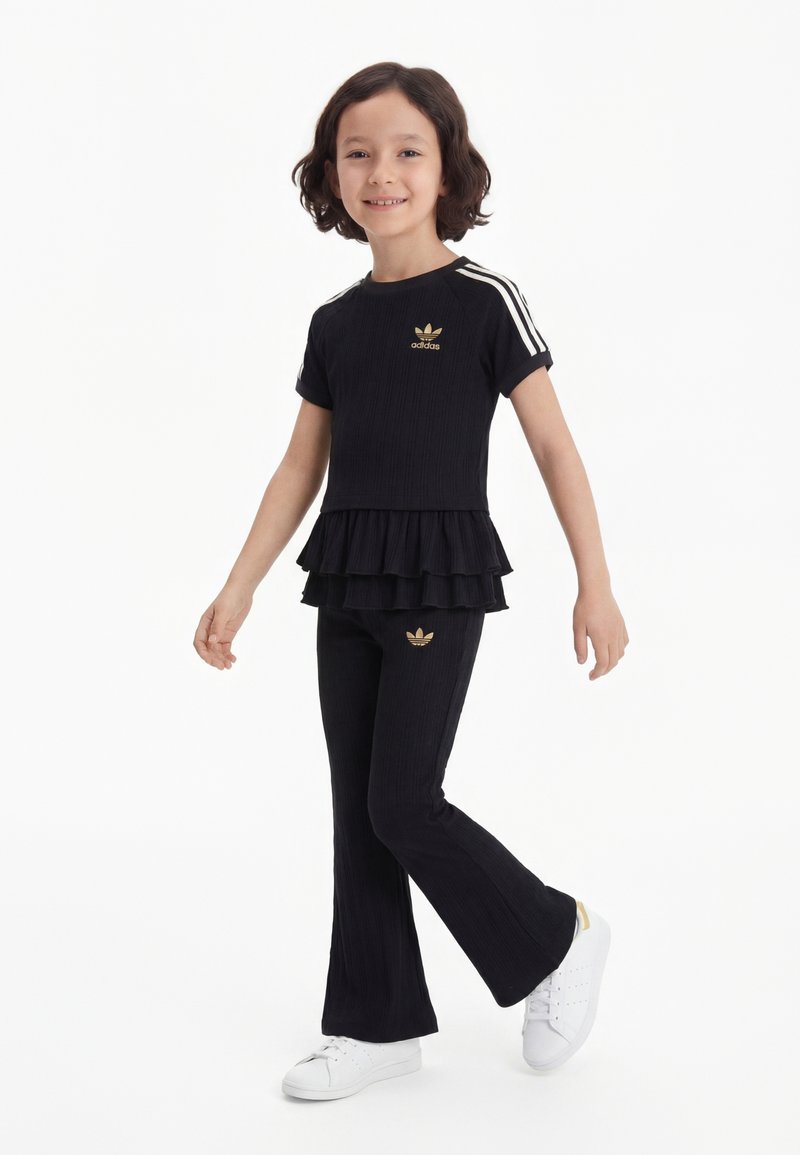 Jeune fille souriante, portant une tenue Adidas noire avec des logos dorés, des bandes blanches sur les manches, un haut noir à volants, un pantalon évasé et des baskets blanches.