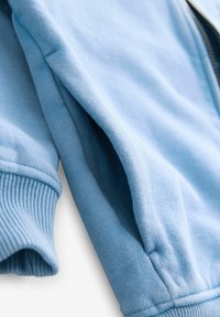 Sudadera azul claro con puños y dobladillo acanalados, que presenta una textura de algodón suave y un cierre de cremallera en la parte frontal.