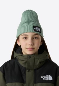 Πλεκτός σκούφος σε ανοιχτό πράσινο, με ραβδωτή υφή και εμφανές μαύρο λογότυπο. Συνδυάζεται με σκούρο πράσινο και μαύρο σακάκι.