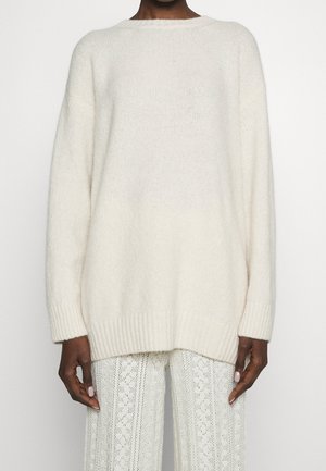 Osoba ubrana w dopasowany, długi biały sweter z dzianiny i białe spodnie z wzorem koronkowym, widoczna od szyi do bioder.