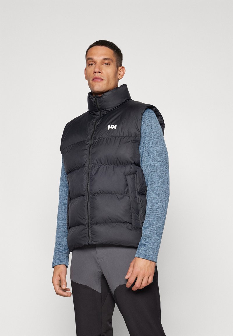 Black padded vest with high collar, front zipper, and logo. Worn over a blue long-sleeve shirt, paired with dark, fitted pants.