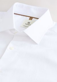Camicia da uomo bianca a vestibilità regolare con colletto appuntito e bottone singolo visibile, etichettata "NEXT" sull'etichetta interna del colletto.
